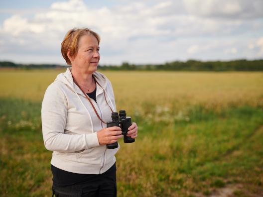 Keski-ikäinen nainen seisoo pellolla, katsoo horisonttiin ja pitelee kiikareita kädessään.