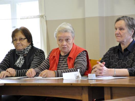 Kolme ikäntynyttä naista istuu pöydän takana ja punaliivinen nainen keskellä puhuu, pöydällä on papereita.