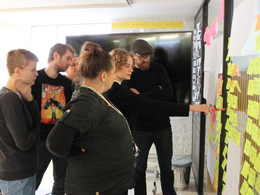 Kuvassa pelisprintin osallistujat katselevat yhdessä tuotettuja, pelin sisältöön liittyviä, ideoita, jotka on kirjattu liimalapuille ja asetettu yhteiselle seinälle, kaikkien näkyville.