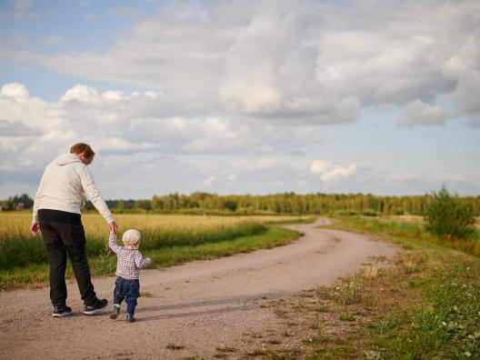 Isovanhempi ja pikkulapsi kävelevät polkua pitkin kauniissa maalaismaisemassa