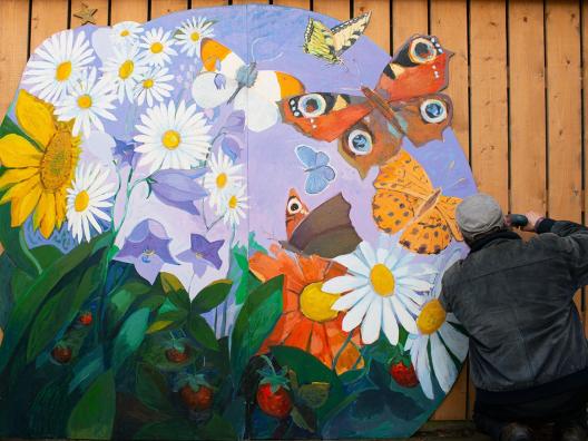 Taitelija Pekka Kainulainen kiinnittää betoniseinään taideteosta, jossa kedon kukkia, erhosia ja runsautta.