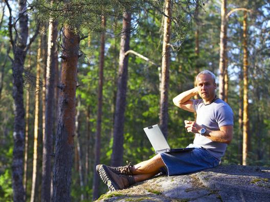 Mies istuu metsän keskellä kalliolla kannettava tietokone sylissä. 