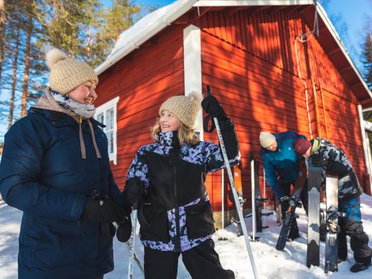 Nainen ja tyttö hymyilevät auringon paisteessa hankien keskellä. Mies ja poika huoltavat liukulumikenkiä taustalla.