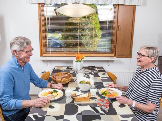 Kaksi ikäihmistä, mies ja nainen, istuvat keittiön pöydän ääressä syömässä ateriaa.