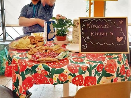 Kuvassa on pöytä, jossa kahvila tarjoiluja. Pöydän takana on lippispäinen mies.