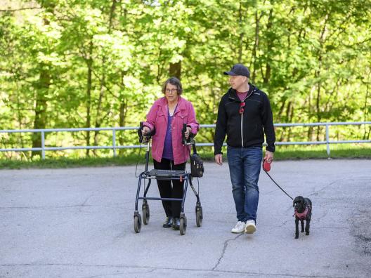 Nuori mies kävelee koiran kanssa rollaattoria käyttävän iäkkään naisen rinnalla