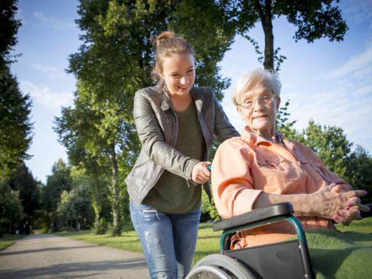 Nainen avustaa ikääntynyttä pyörätuolilla ulkoilussa.