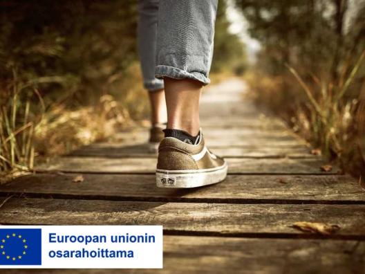 Ihminen kävelee puisella polulla, vain jalat näkyvät, alareunassa sininen EU-lippulogo ja teksti jossa lukee: Euroopan unionin osarahoittama