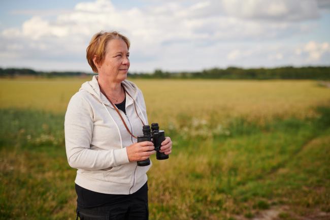 Keski-ikäinen nainen seisoo pellolla, katsoo horisonttiin ja pitelee kiikareita kädessään.