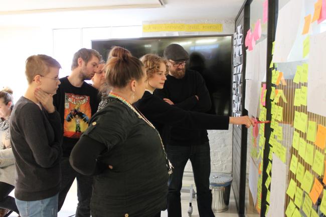 Kuvassa pelisprintin osallistujat katselevat yhdessä tuotettuja, pelin sisältöön liittyviä, ideoita, jotka on kirjattu liimalapuille ja asetettu yhteiselle seinälle, kaikkien näkyville.