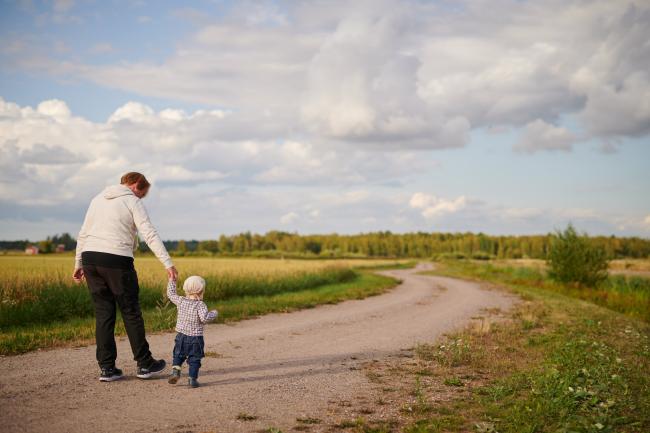 Isovanhempi ja pikkulapsi kävelevät polkua pitkin kauniissa maalaismaisemassa