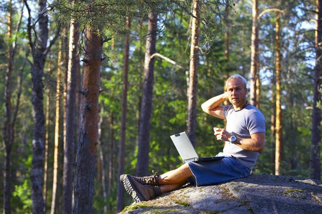 Mies istuu metsän keskellä kalliolla kannettava tietokone sylissä. 