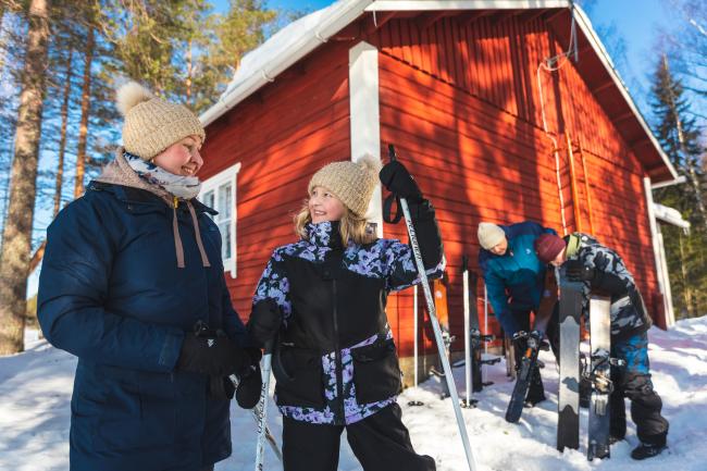 Nainen ja tyttö hymyilevät auringon paisteessa hankien keskellä. Mies ja poika huoltavat liukulumikenkiä taustalla.