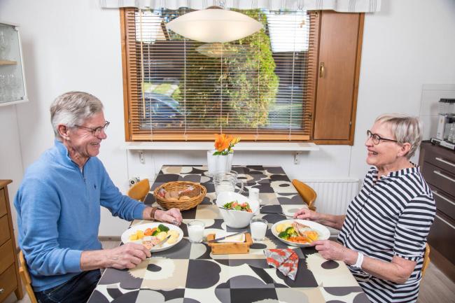 Kaksi ikäihmistä, mies ja nainen, istuvat keittiön pöydän ääressä syömässä ateriaa.