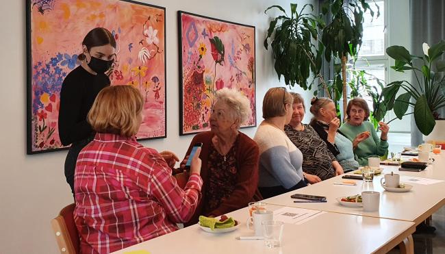 Kuusi naista istuu pöydän ääressä ja yksi nainen seisoo. Naiset keskustelevat keskenään.