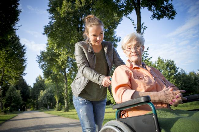 Nainen avustaa ikääntynyttä pyörätuolilla ulkoilussa.