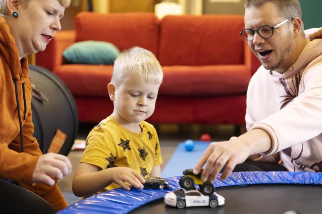 Oranssipaitainen äiti, keltapaitainen pikkupoika ja isä, jolla vaaleanpunainen Pikku Myy -huppari,  leikkivät autoilla perheiden kohtaamispaikassa.