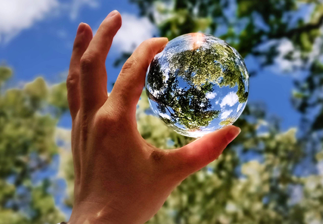 Kohti puidenlatvoja ja pilviä kohotettu käsi, jossa peukalon ja etusormen välissä oleva lasipallo toistaa näkymää.